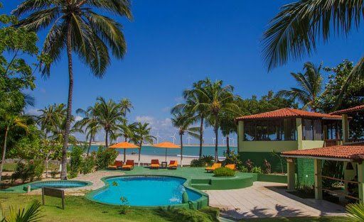 Uma piscina rodeada por palmeiras, espreguiçadeiras e guarda-sóis em um resort à beira-mar sob um céu azul brilhante.