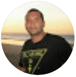 Uma pessoa vestindo uma camiseta preta estampada sorri em uma praia durante o pôr do sol.