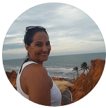 Uma pessoa sorridente posando com uma regata branca, tendo como pano de fundo uma praia, o oceano e falésias alaranjadas.