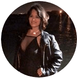 Uma pessoa vestindo jaqueta e blusa pretas sorri, posando ao ar livre à noite com reflexos de luz escuros e desfocados ao fundo.