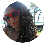 Uma mulher com longos cabelos cacheados usa óculos de sol vermelhos em formato de lábios, fotografada ao ar livre perto de palmeiras e água.