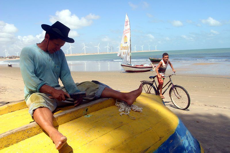 Uma pessoa de chapéu está sentada em um barco amarelo em uma praia ensolarada, trabalhando com corda, enquanto outra pessoa está ao lado de uma bicicleta.