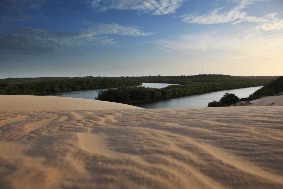 Dunas de areia em primeiro plano oferecem vista para um rio que serpenteia por uma paisagem verdejante sob um céu azul ao pôr do sol.