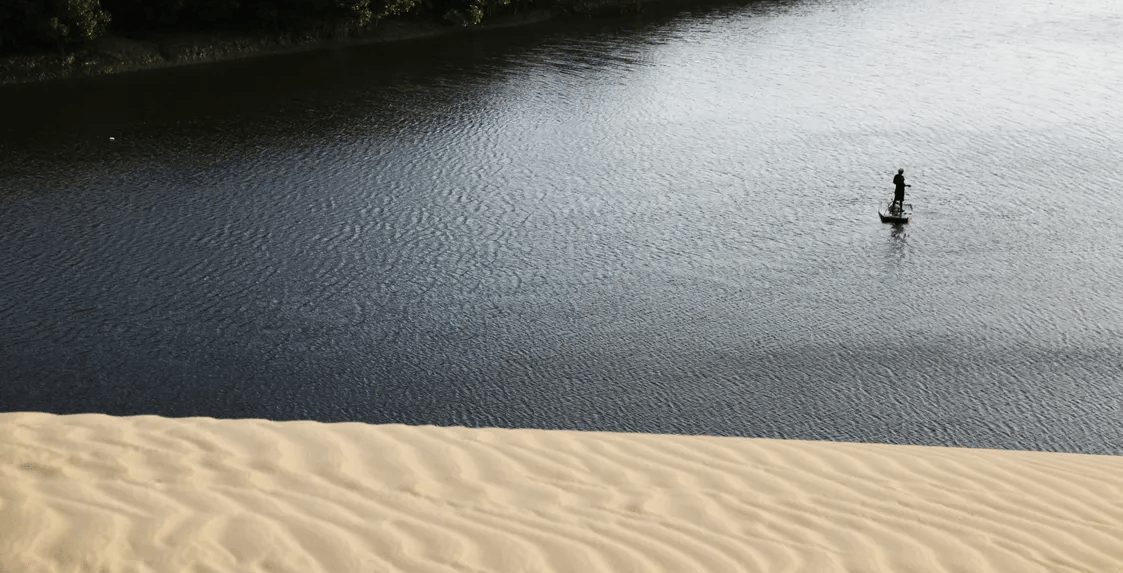Uma figura solitária pratica stand-up paddle em águas escuras e ondulantes, vista do topo de uma duna de areia texturizada.
