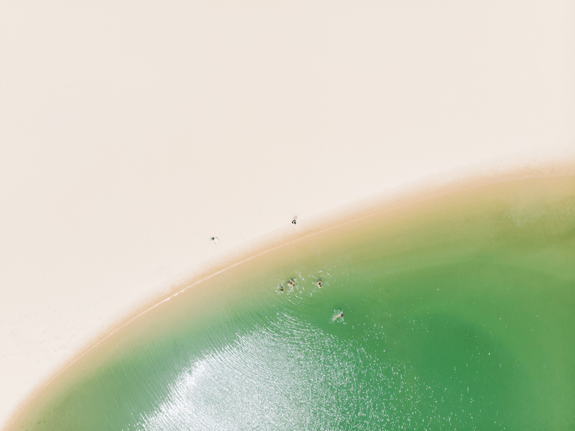 Vista aérea da areia branca encontrando a água rasa e vibrante de cor verde, com algumas pequenas figuras visíveis na costa.
