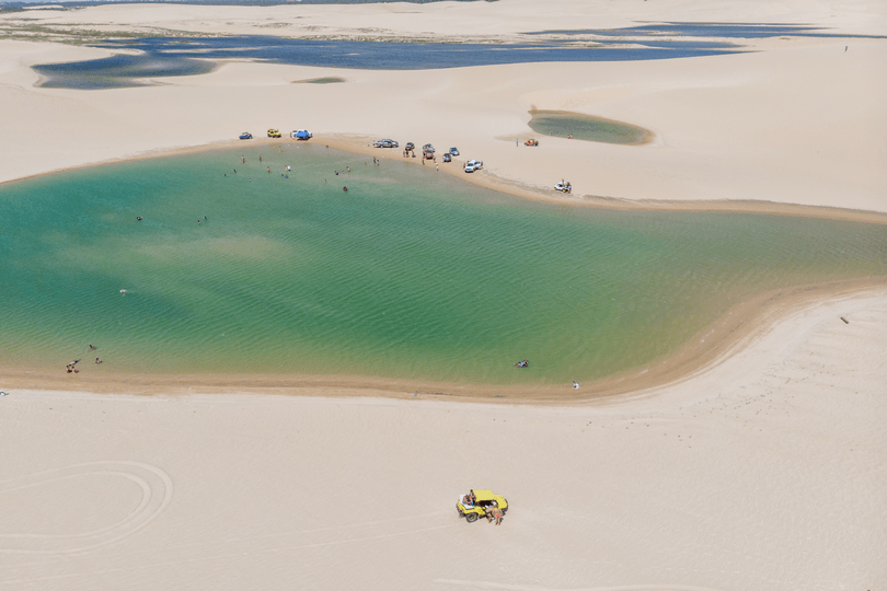 Vista aérea de uma lagoa turquesa rodeada por vastas dunas de areia clara, com veículos estacionados e pessoas nadando.