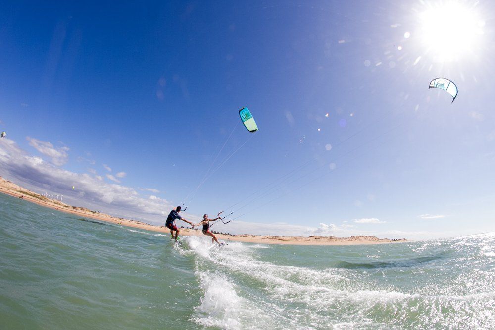 Duas pessoas praticando kitesurf no oceano sob um céu claro e ensolarado, com dunas de areia ao fundo.