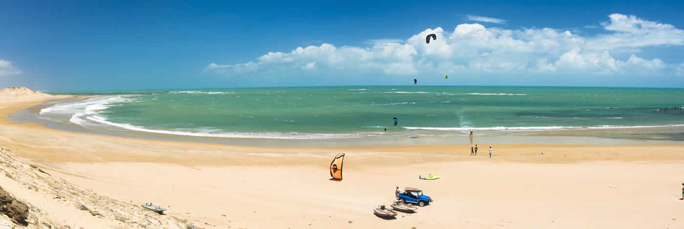 Uma vista panorâmica de uma praia de areia com água turquesa, um kitesurfista ao longe e veículos estacionados na costa.