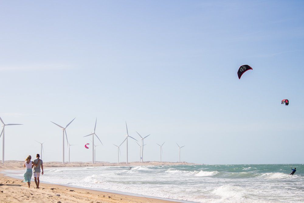 Um casal caminha por uma praia de areia perto de turbinas eólicas, enquanto kitesurfistas deslizam no oceano sob um céu azul límpido.