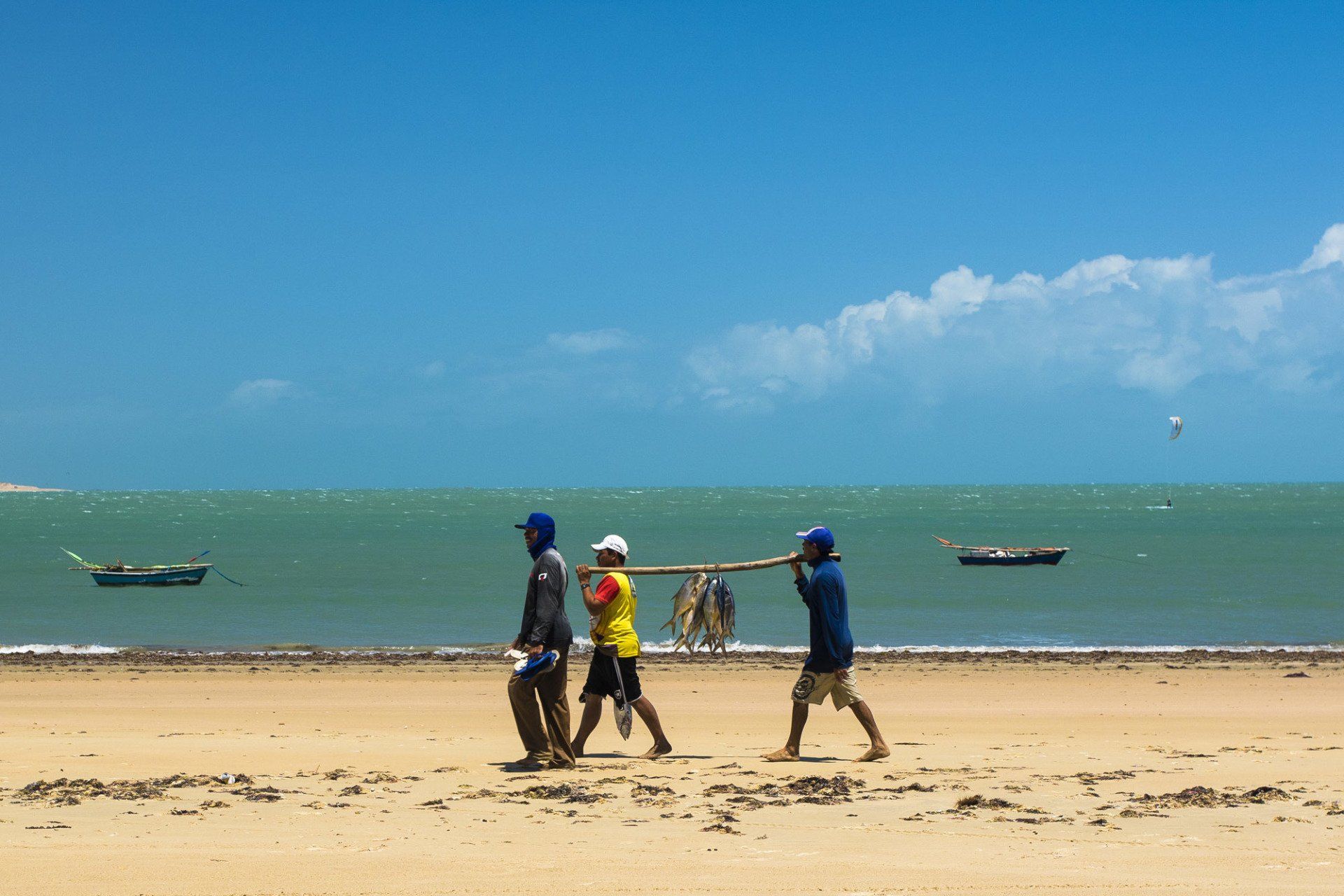 Três pessoas carregam uma longa vara através de uma praia arenosa, com pequenos barcos flutuando no oceano ao fundo.