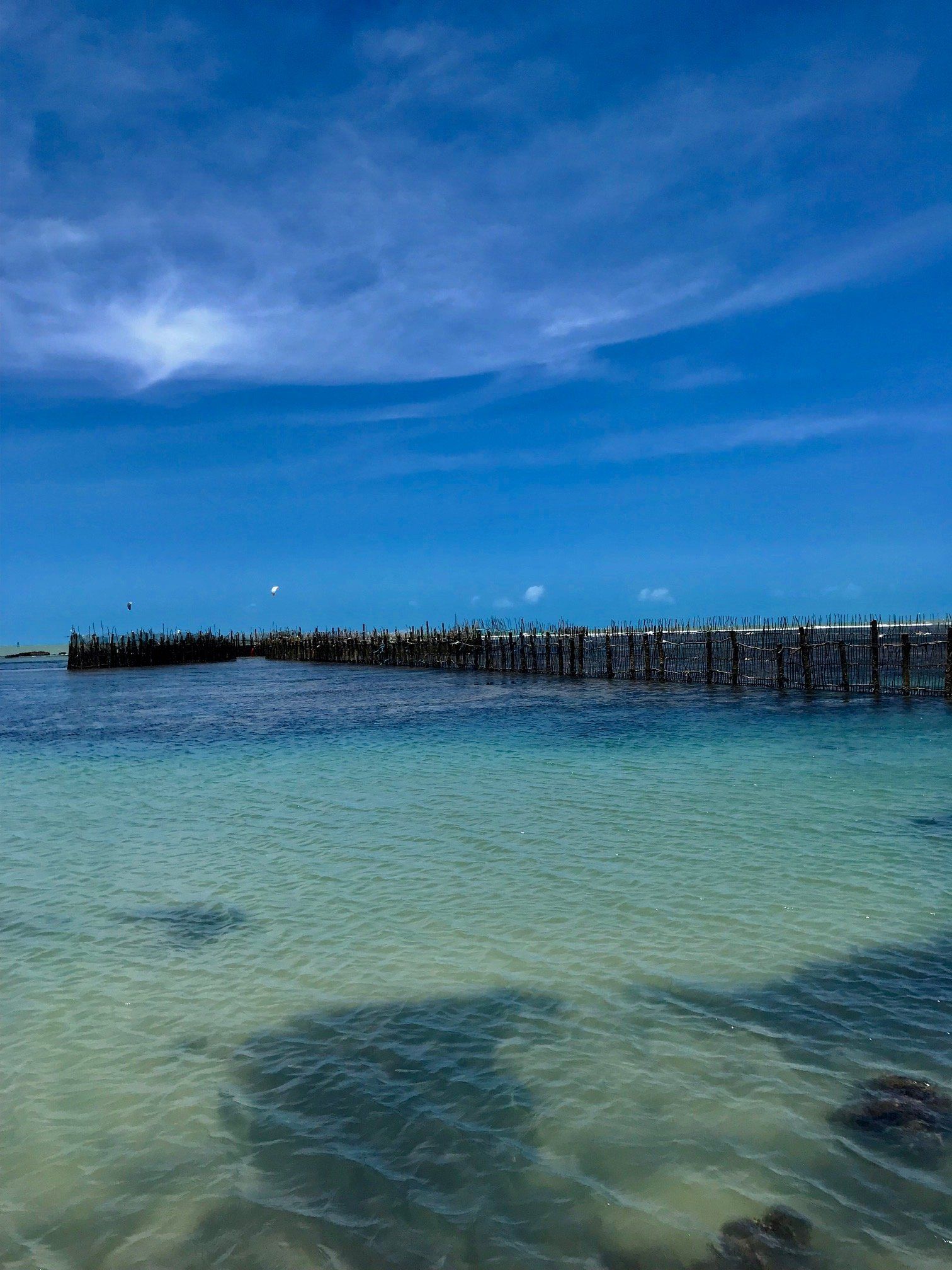 Um mar azul-turquesa raso conduz a um longo cais de madeira desgastado pelo tempo, sob um céu azul profundo com nuvens finas e esparsas.
