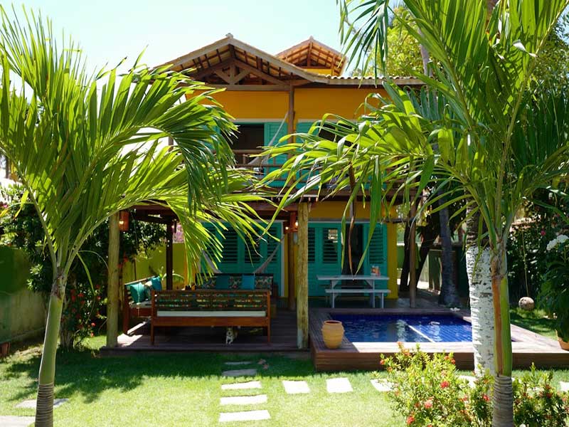 Uma casa amarela de dois andares com persianas verde-azuladas, um deck de madeira, móveis de jardim e uma pequena piscina rodeada por palmeiras.