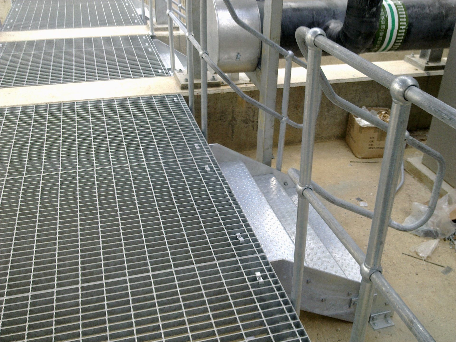 Steel grating walkway with handrails and step leading to industrial equipment.