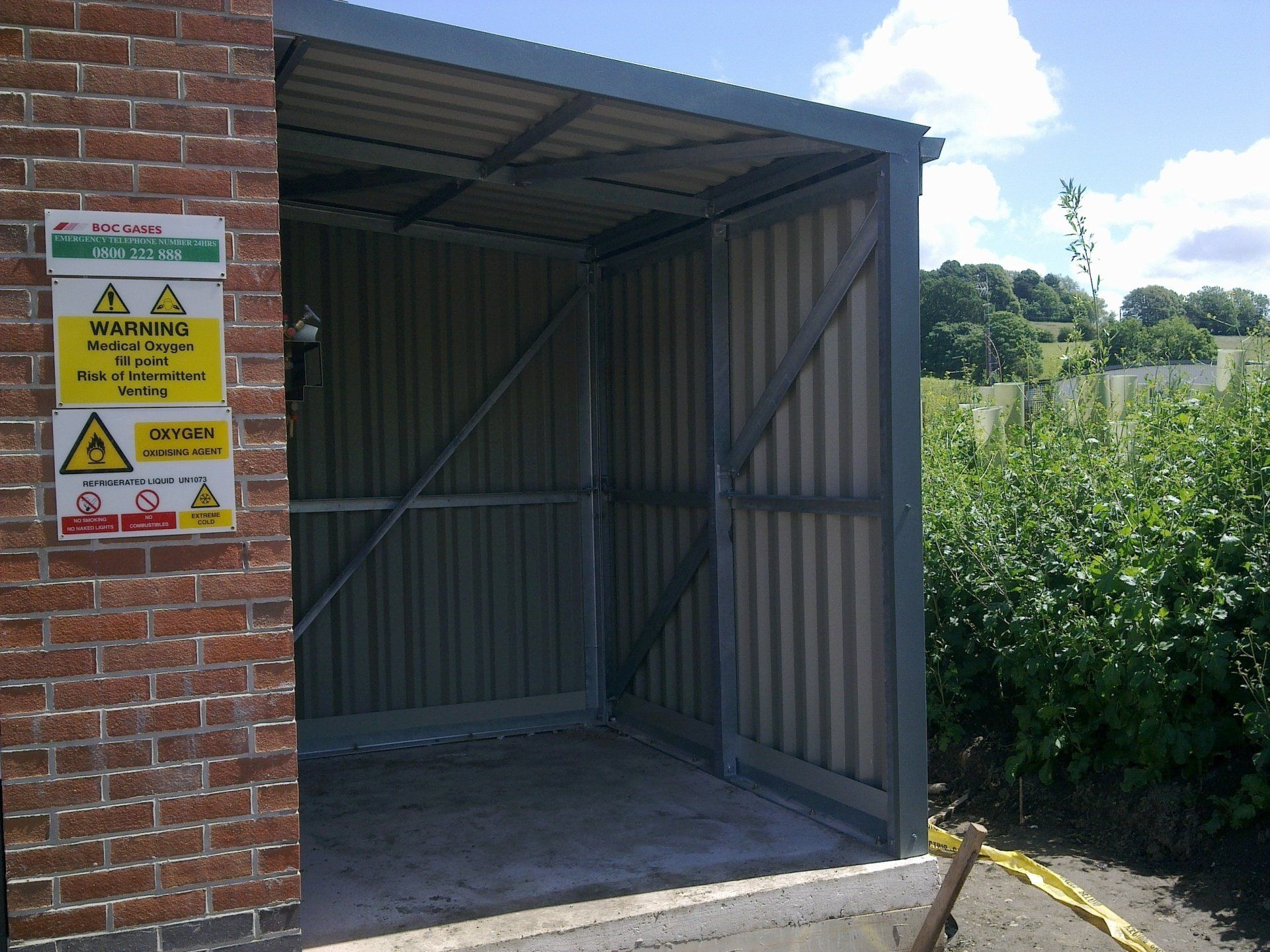 Metal shed next to a brick wall with warning signs. Open entrance, corrugated steel walls and roof.