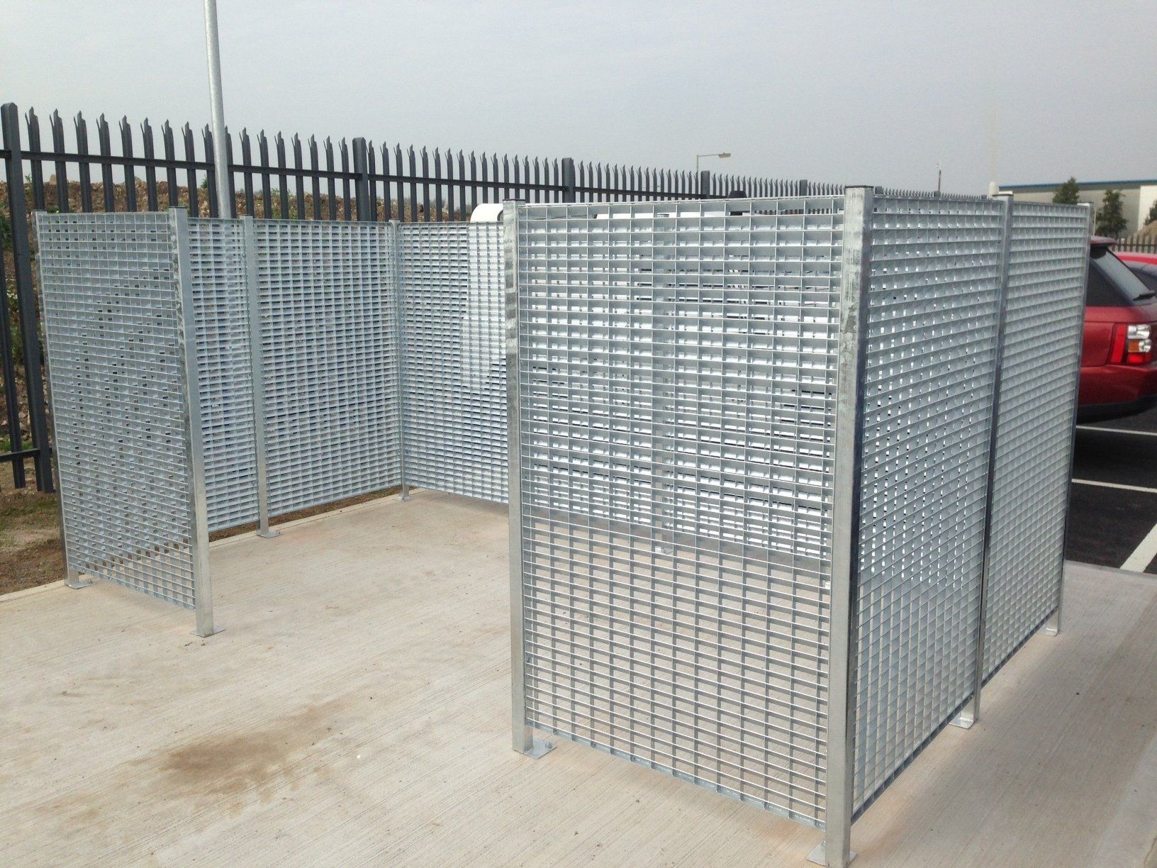 Metal mesh enclosure, possibly for waste disposal, in a paved area, with a fence and car in background.
