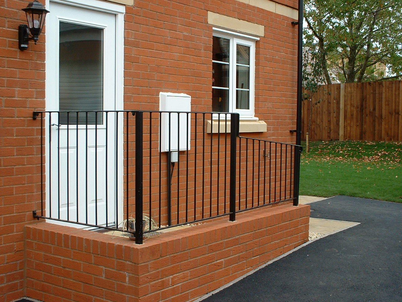 Brick building with white door, window, and black railing. Asphalt path and grass lawn in the setting.