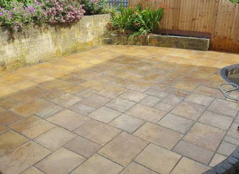 Light coloured stone patio with a stone wall and wooden fencing next to it with flowers and plants
