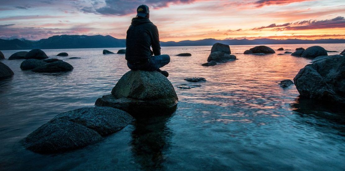 A man is sitting on a rock overlooking the ocean at sunset.
