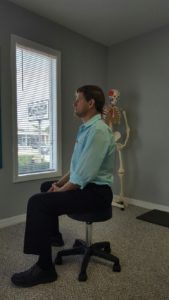 A man is sitting on a stool in a room with a skeleton in the background.