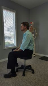 A man is sitting on a chair in a room with a skeleton in the background.