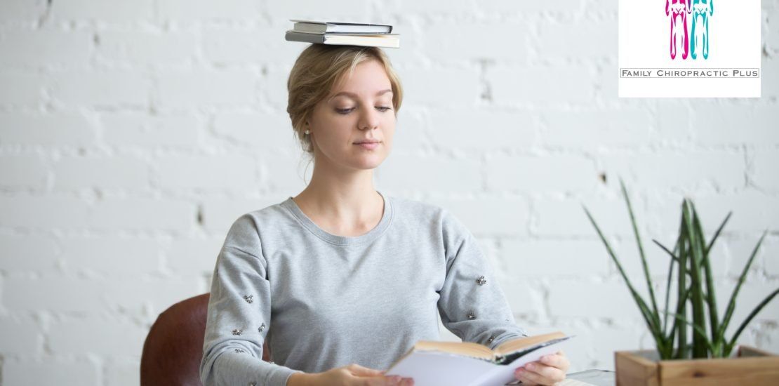 A woman is sitting at a desk with a stack of books on her head.