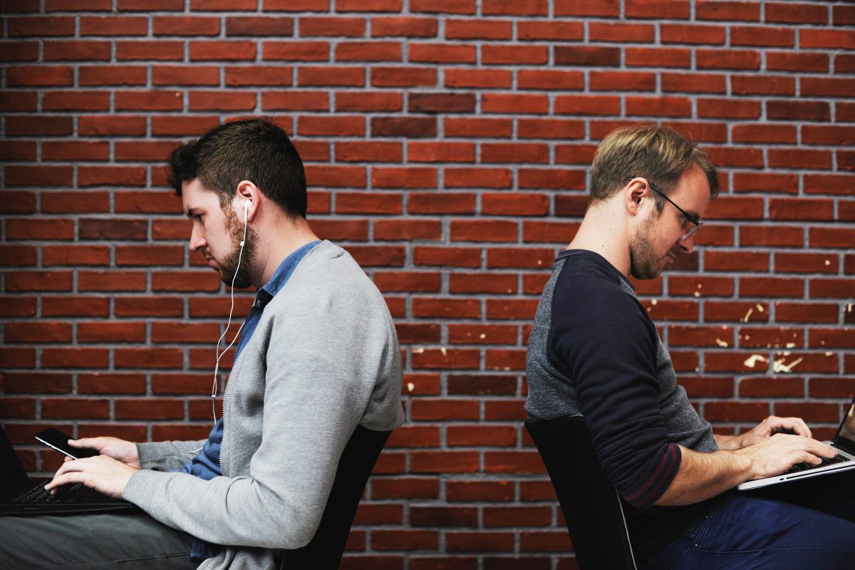 Two men are sitting back to back using laptops in front of a brick wall.