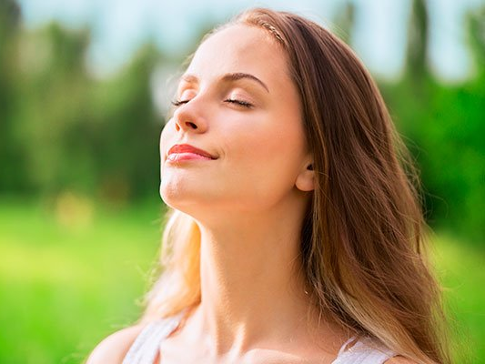A woman is breathing fresh air in a park with her eyes closed.