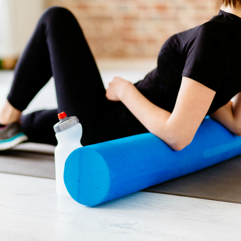 A woman is laying on a yoga mat using a blue foam roller