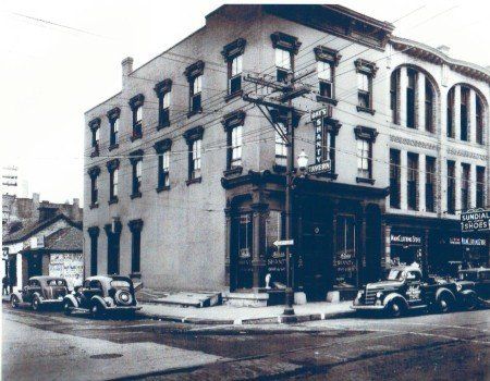 A historic three-story brick corner building with arched windows, vintage cars parked out front, and a prominent sign.