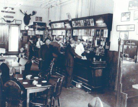 A black-and-white photo shows a bustling 19th-century saloon with patrons at a bar and tables, and a moose head on the wall.