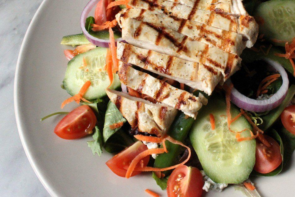 A grilled chicken salad on a light plate, featuring sliced chicken, tomatoes, cucumber slices, and red onion rings.