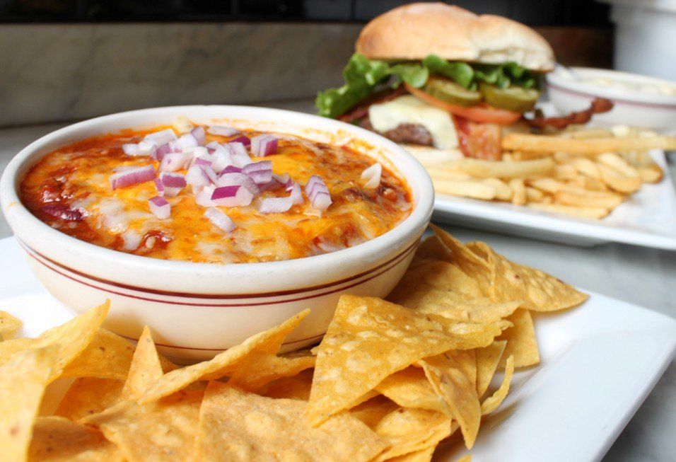 A bowl of chili topped with cheese and red onions, served with tortilla chips, beside a burger and fries on a table.