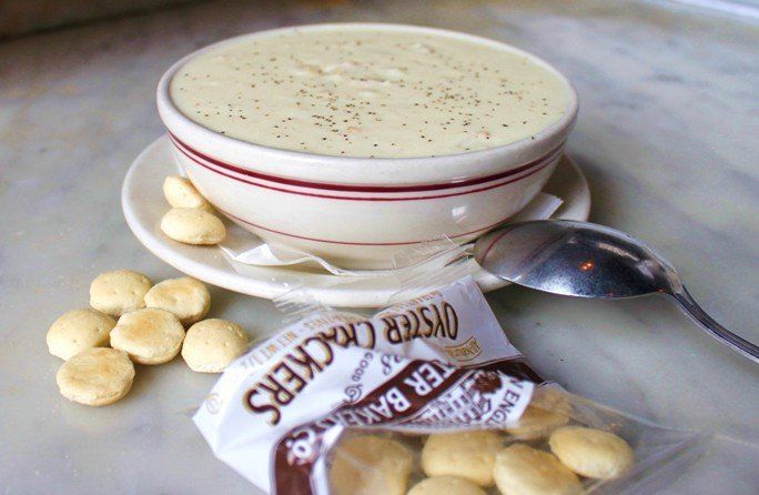 A bowl of creamy chowder with black pepper, served on a white plate with loose and packaged oyster crackers and a spoon.