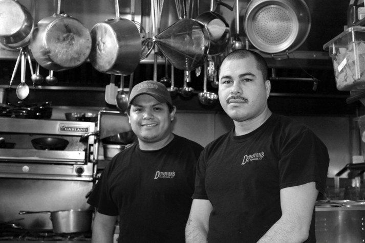 Two chefs wearing black restaurant uniforms stand in a professional kitchen with hanging cookware in the background.