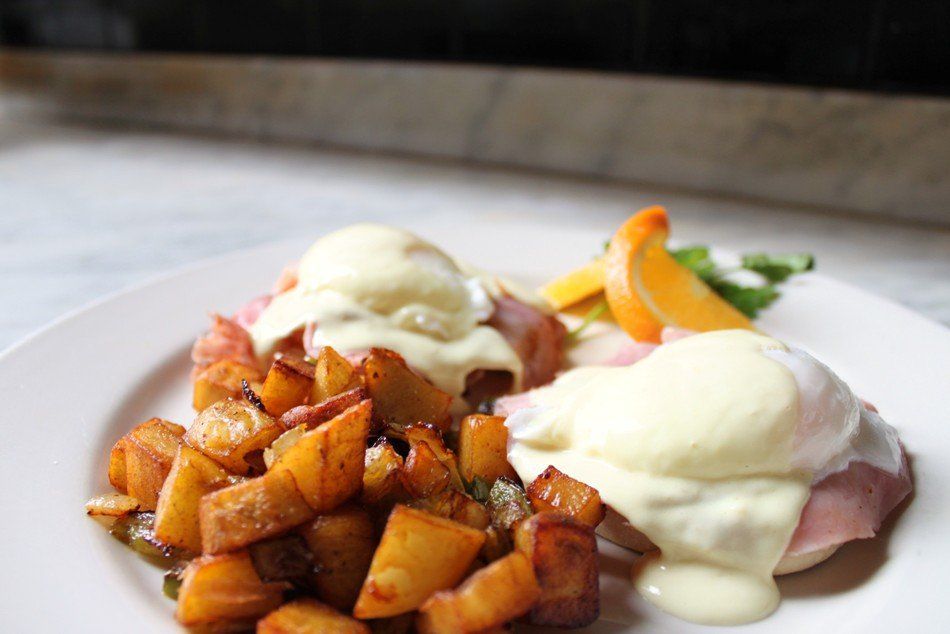 A plate of Eggs Benedict with hollandaise sauce, sliced ham, and a generous side of crispy home-style breakfast potatoes.