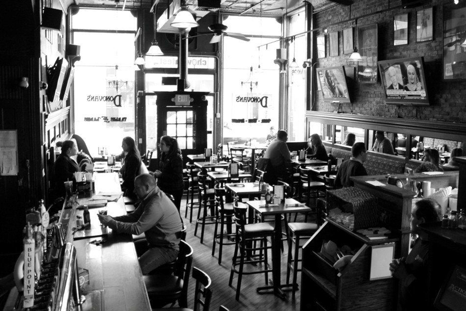 A black-and-white interior shot of a bar with patrons sitting at the counter and at small tables.