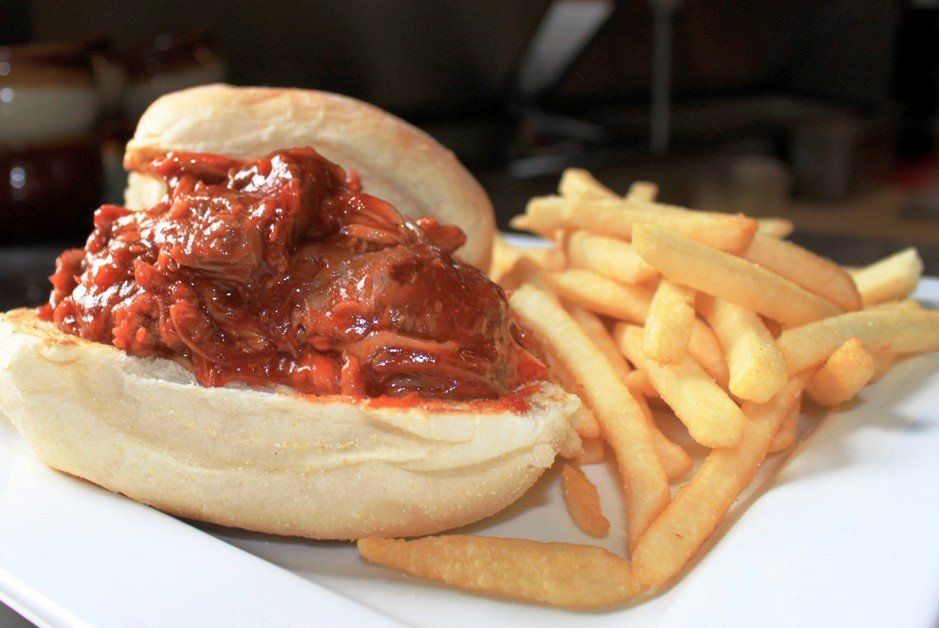 A barbecue beef sandwich topped with thick, reddish-brown sauce, served with golden French fries on a white plate.