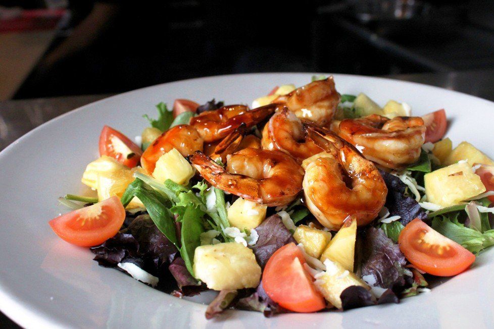 A bowl of salad topped with glazed grilled shrimp, pineapple chunks, and tomato wedges on a white plate.