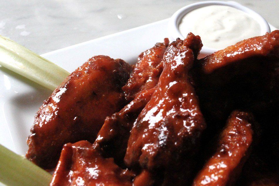 A plate of glazed, red buffalo wings served with celery sticks and a side of white dipping sauce.