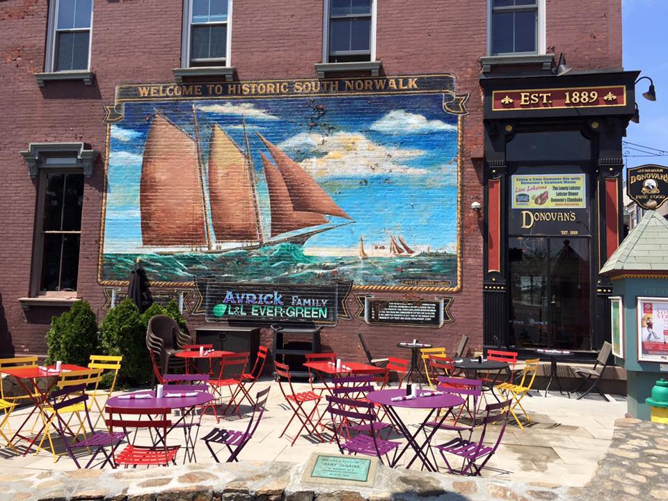 Outdoor seating with colorful cafe tables and chairs in front of a building featuring a nautical mural in South Norwalk.