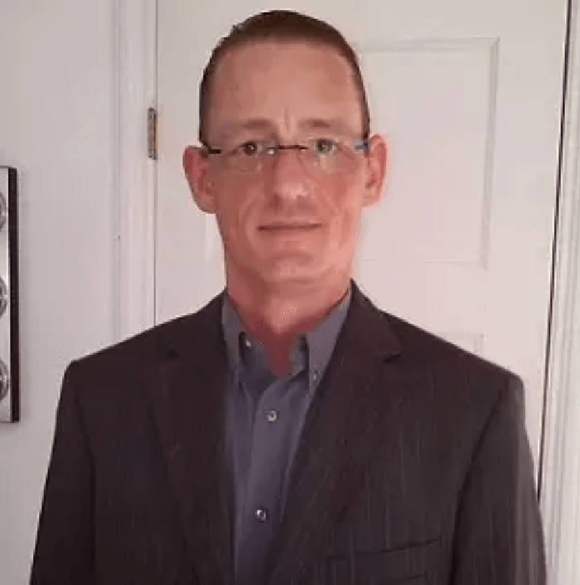 Man in glasses and suit jacket, standing in front of a white door.