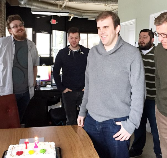 A group of men standing around a cake with candles on it