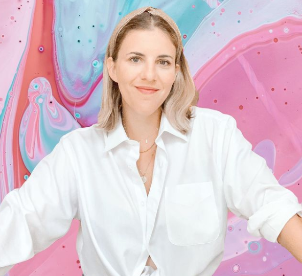 A woman in a white shirt is standing in front of a pink background.