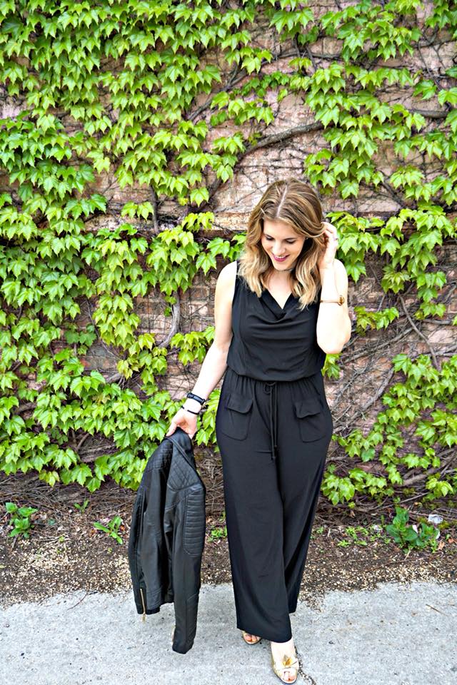 A woman in a black jumpsuit is walking down the street holding a leather jacket.