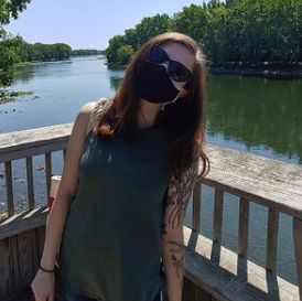A woman wearing a mask and sunglasses is standing on a bridge overlooking a river.