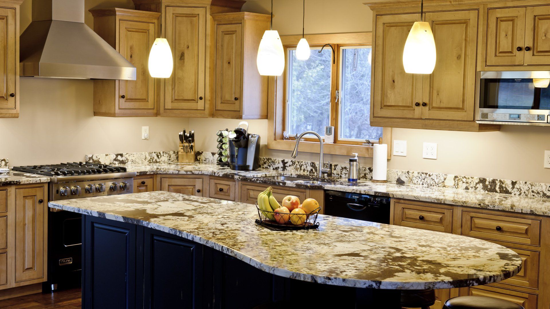 Warm kitchen with wood cabinets, granite island, hanging pendant lights, and a bowl of fruit.
