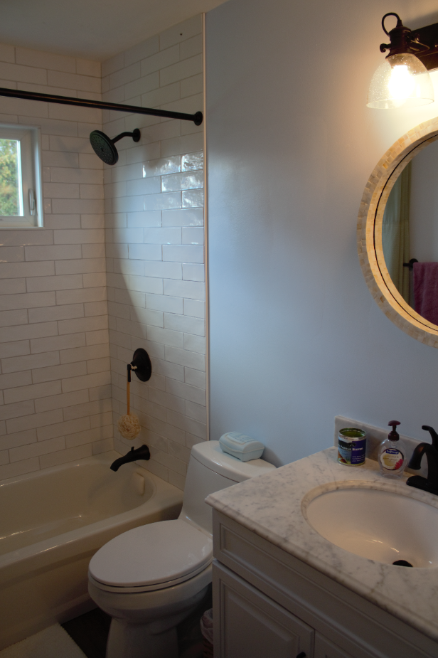 Small bathroom with shower-tub, white tile, vanity sink, and round mirror above counter.