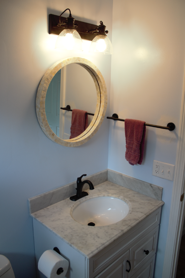 Bathroom vanity with round mirror, black sconce, marble sink, and towel bars on light walls