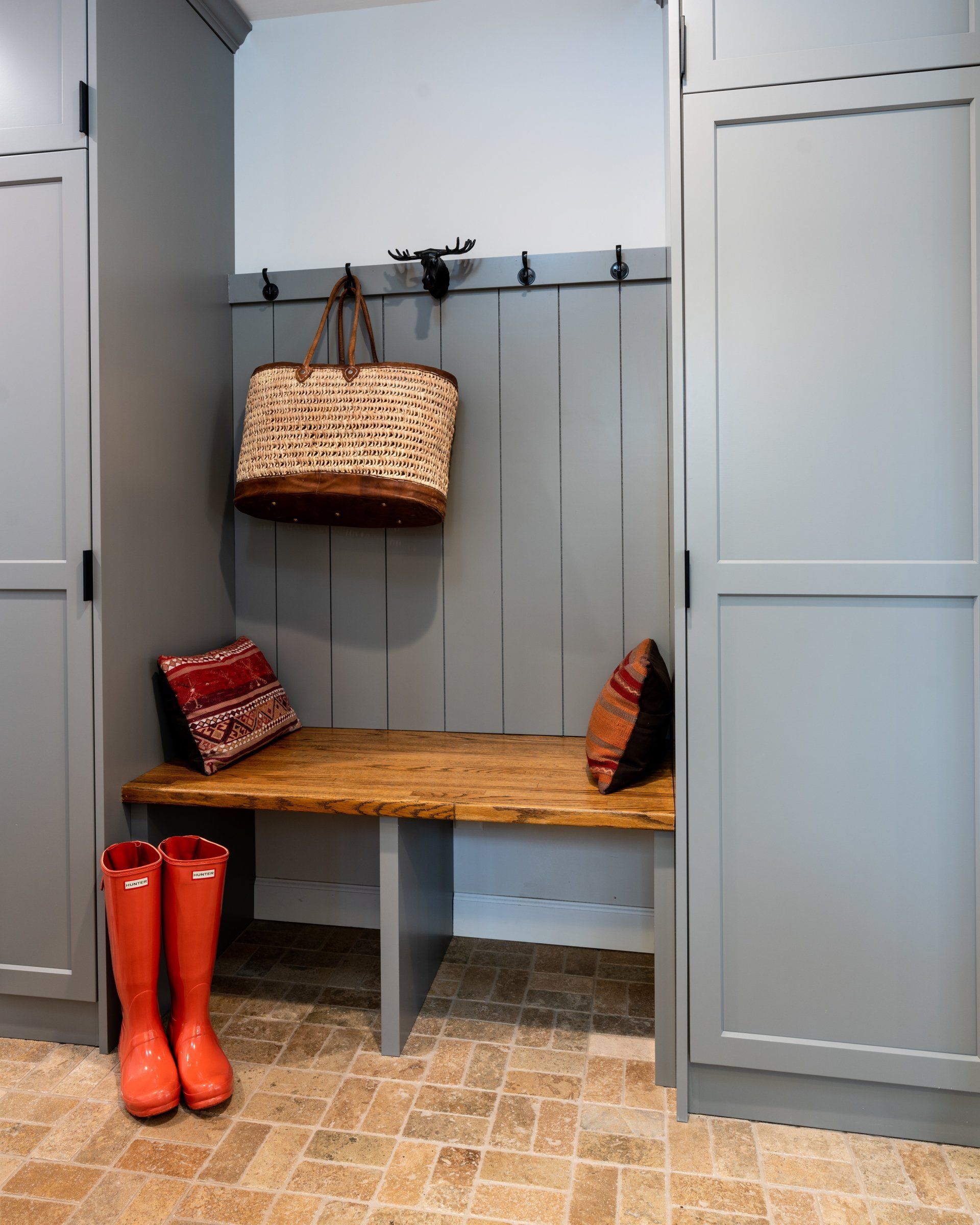 Gray locker-room bench with hanging wicker basket, red boots, and patterned cushions on a tiled floor