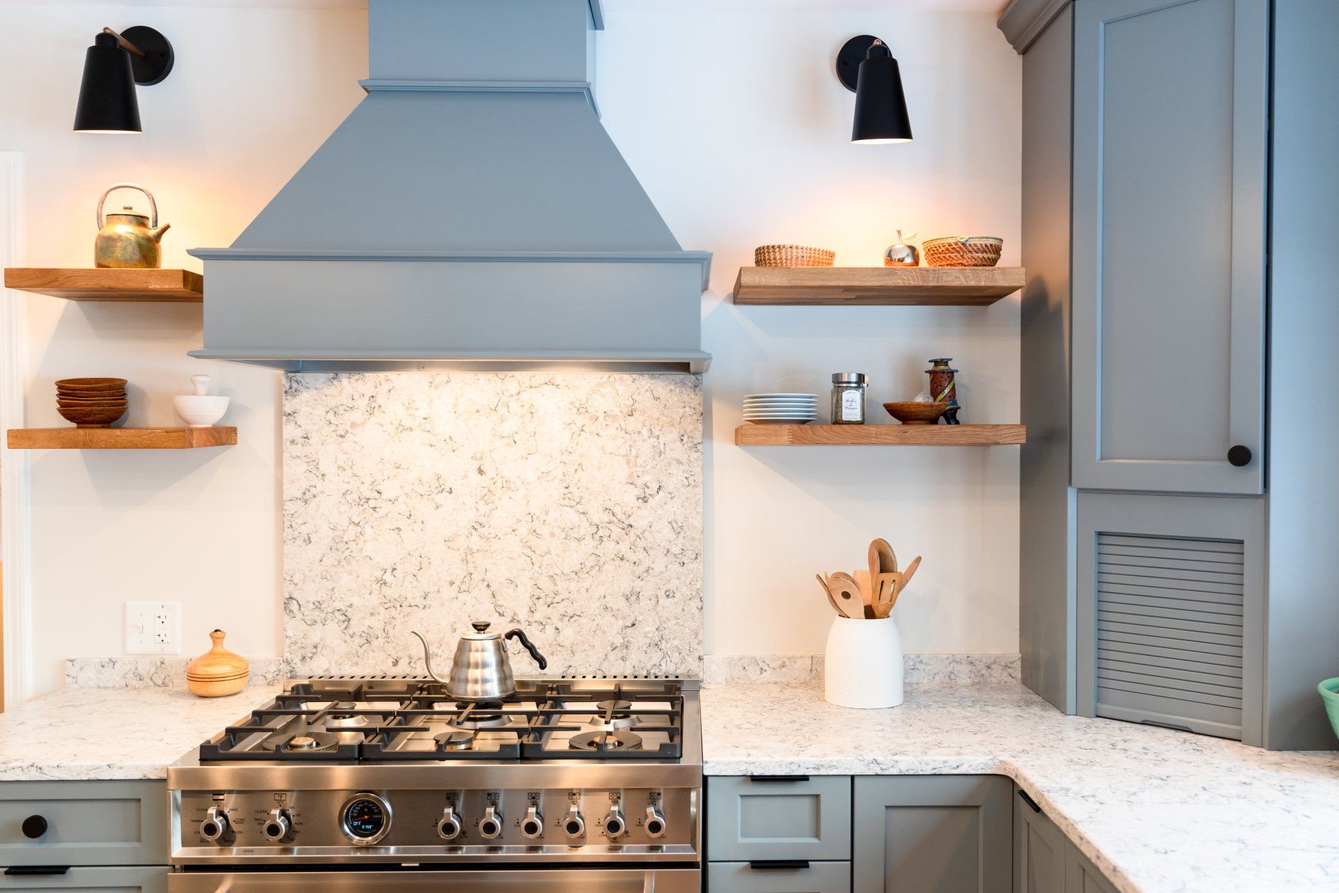 Modern kitchen with stainless stove, gray hood, white countertops, shelves, and black wall sconces